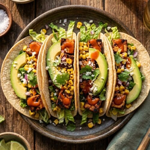 overhead view of BBQ tofu tacos arranged on a rustic wooden table, golden hour lighting, vibrant orange-red bbq sauce drizzle, creamy avocado slices, yellow corn kernels, and crisp green romaine shreds.